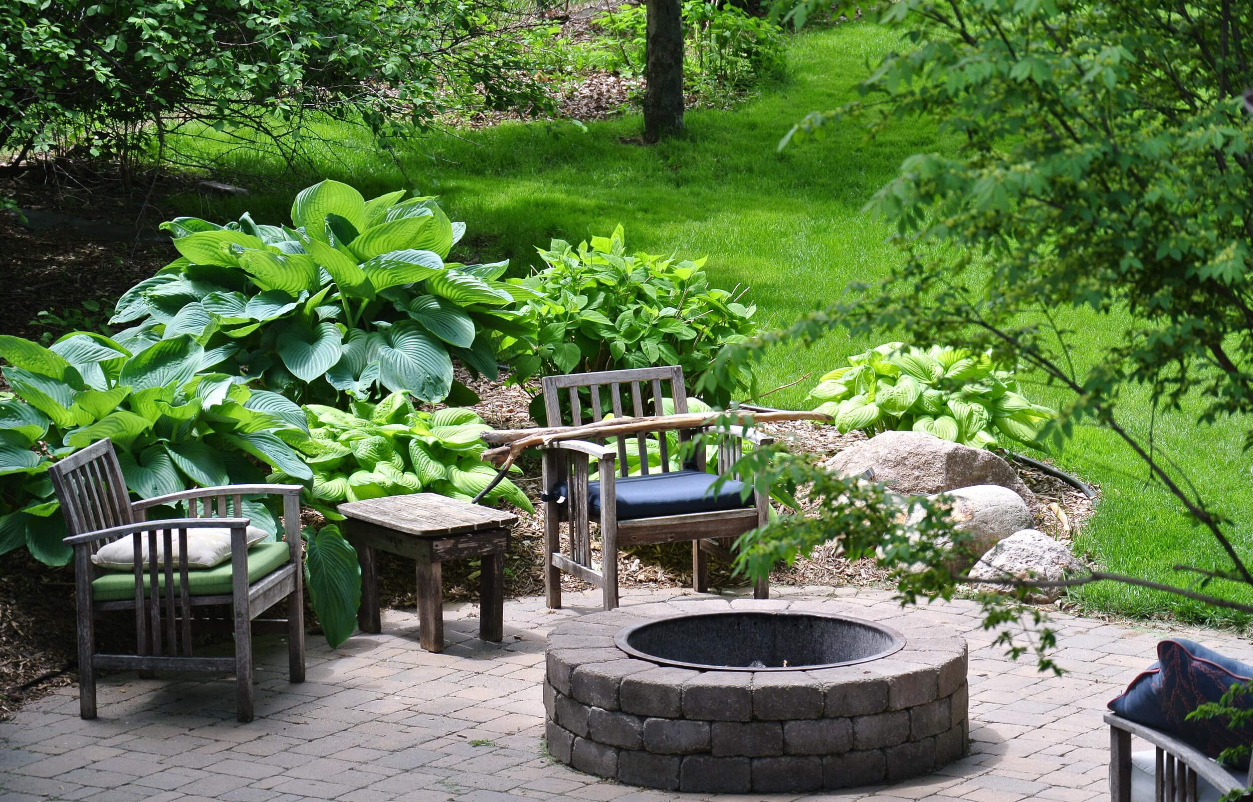 Grüner Garten mit Holzsesseln um eine runde Feuerstelle